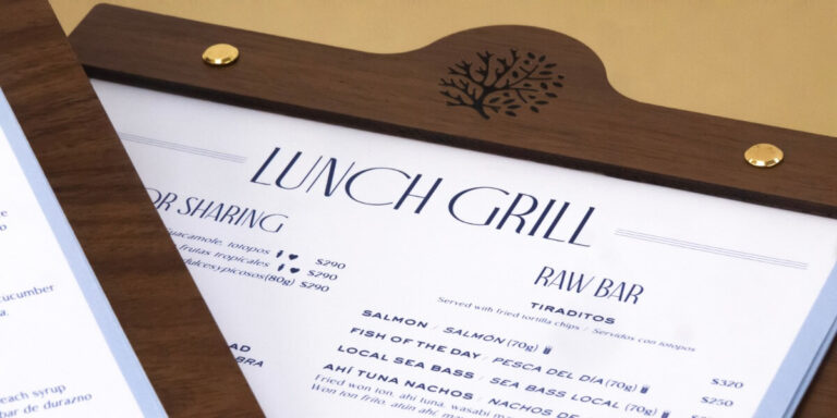 Imagen de un portamenú de madera con detalles metálicos dorados y diseño grabado de coral, mostrando carta de restaurante ‘Lunch Grill’ impresa en papel blanco con tipografía azul elegante, ideal para hoteles y gastronomía en Cancún.