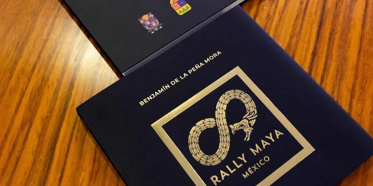 Imagen de libro con portada negra y estampado dorado del Rally Maya México, acompañado del nombre Benjamín de la Peña Mora en letras doradas, colocado sobre mesa de madera junto a otro ejemplar, ejemplo de impresión editorial premium realizada en Cancún por Grupo Regio.